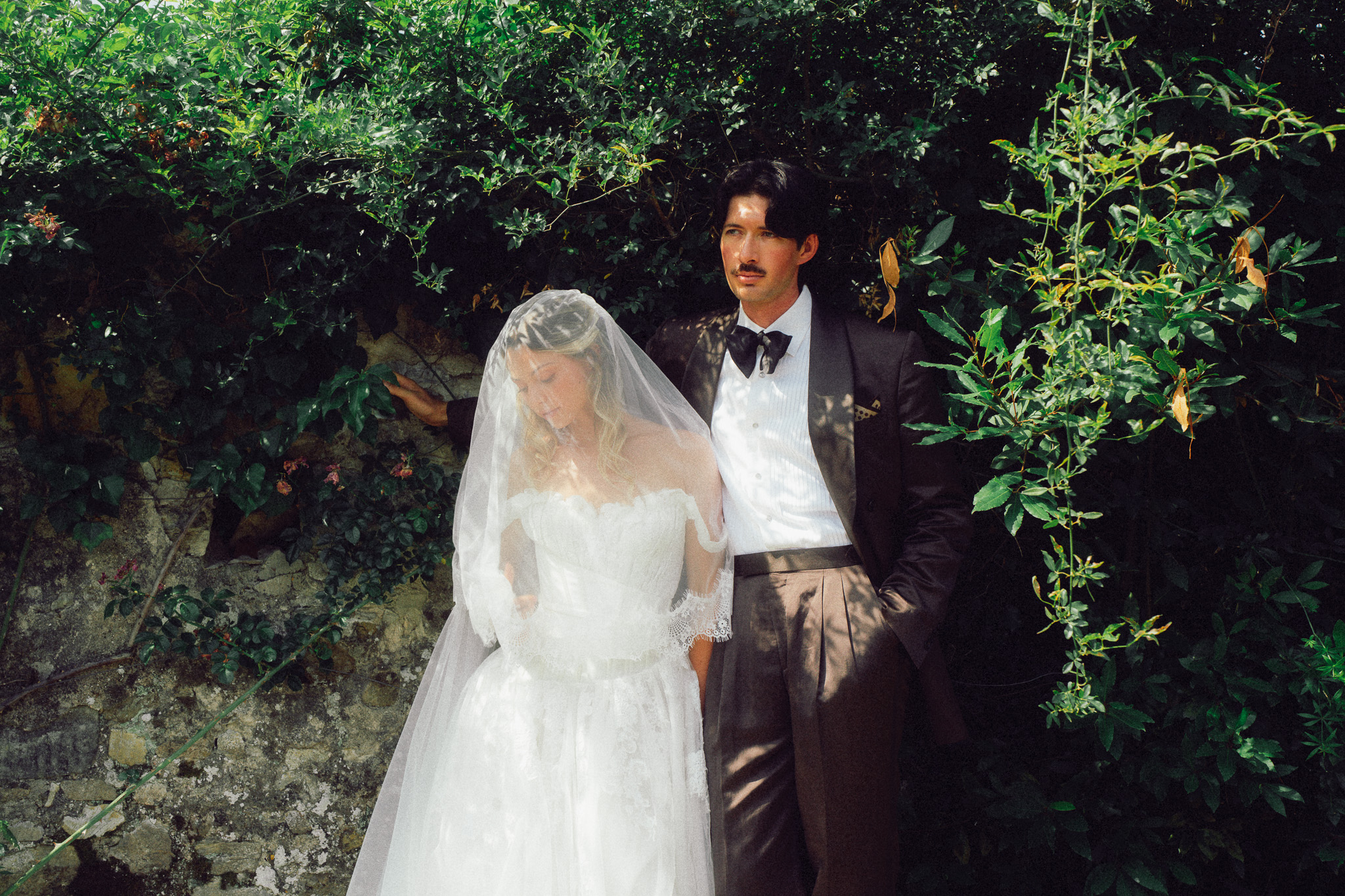 Bride in a classic white wedding gown with a long veil standing beside the groom in a dark tuxedo with a bow tie, posing outdoors against a backdrop of greenery.