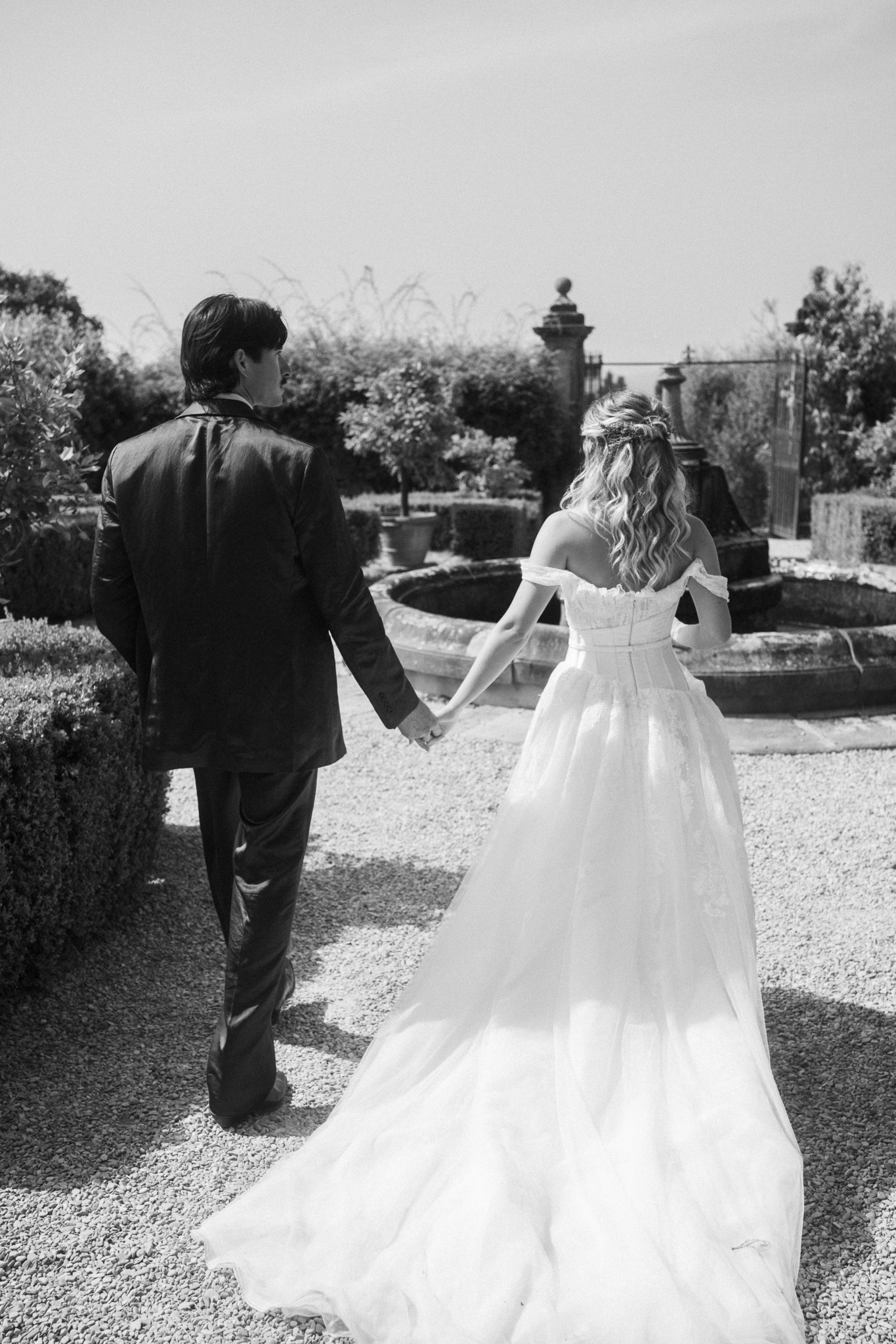 Bride and groom holding hands, walking down a garden path, view from behind.