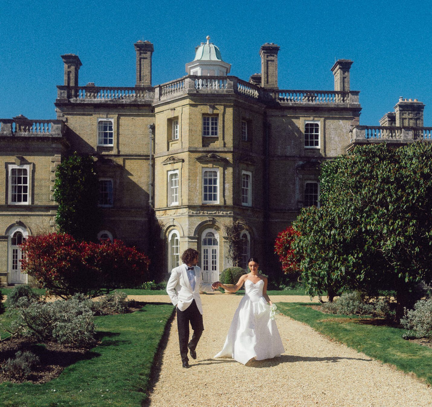 Bride and groom walking hand in hand outside a grand historic manor house under a clear blue sky.