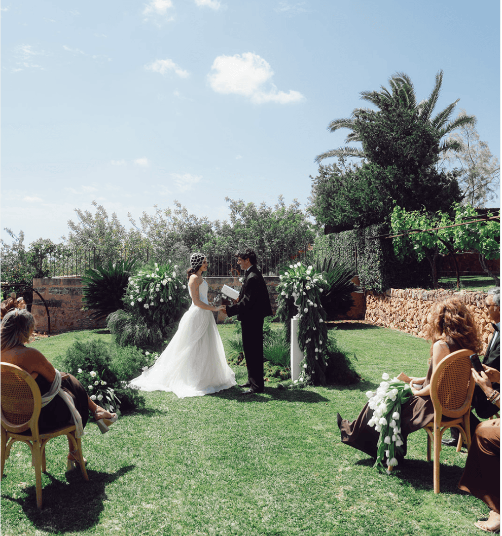 Outdoor Mallorca wedding ceremony with bride and groom surrounded by guests in a sunlit Mediterranean garden.